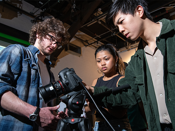 Three students crowd around and examine a piece of camera equipment.